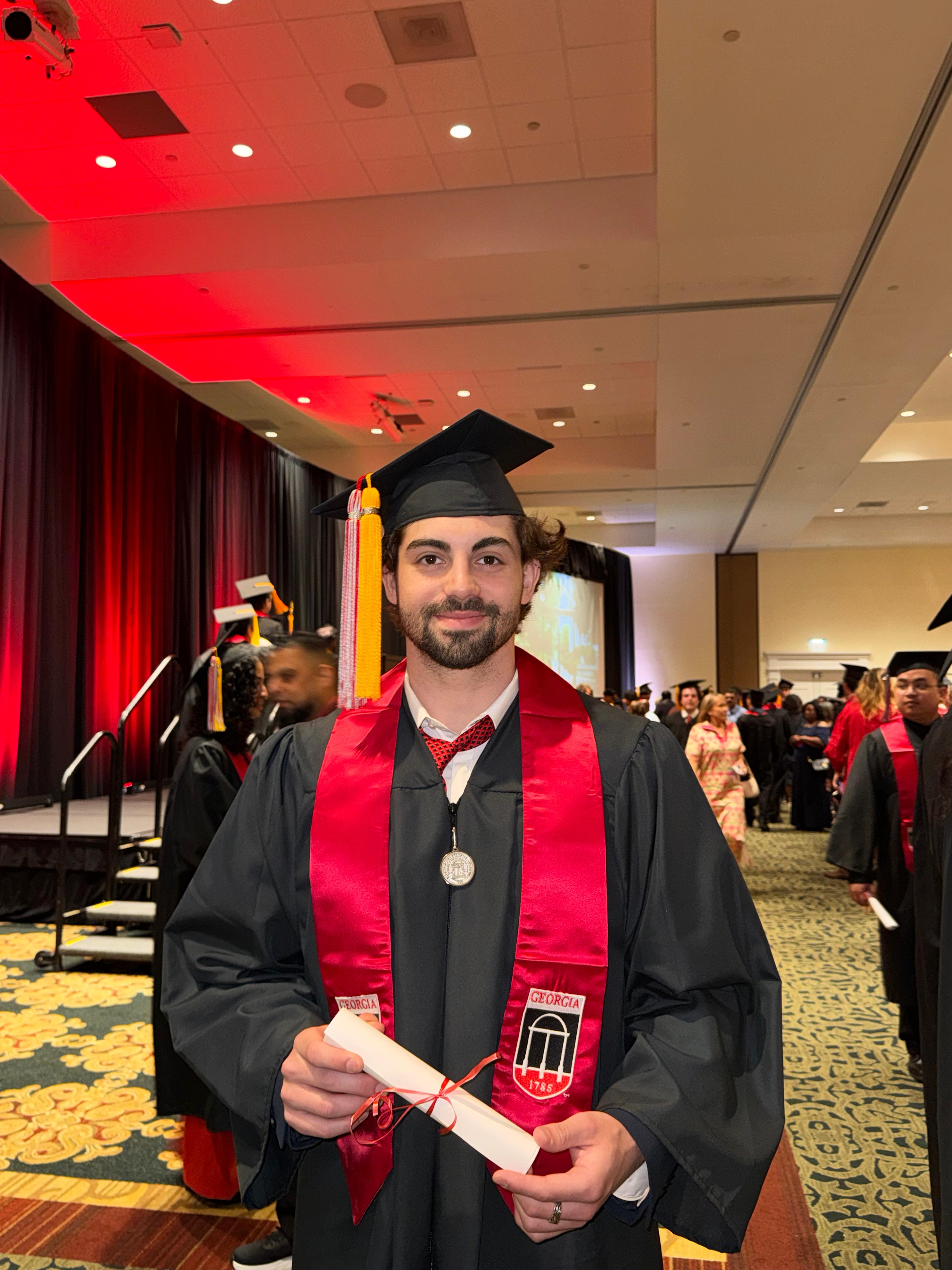 Kristian Pitshugin at graduation in cap and gown holding his diploma.